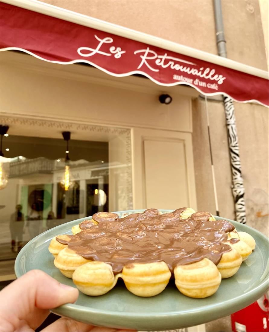 Une gaufre au chocolat tenue devant la façade des Retrouvailles et son store bordeaux