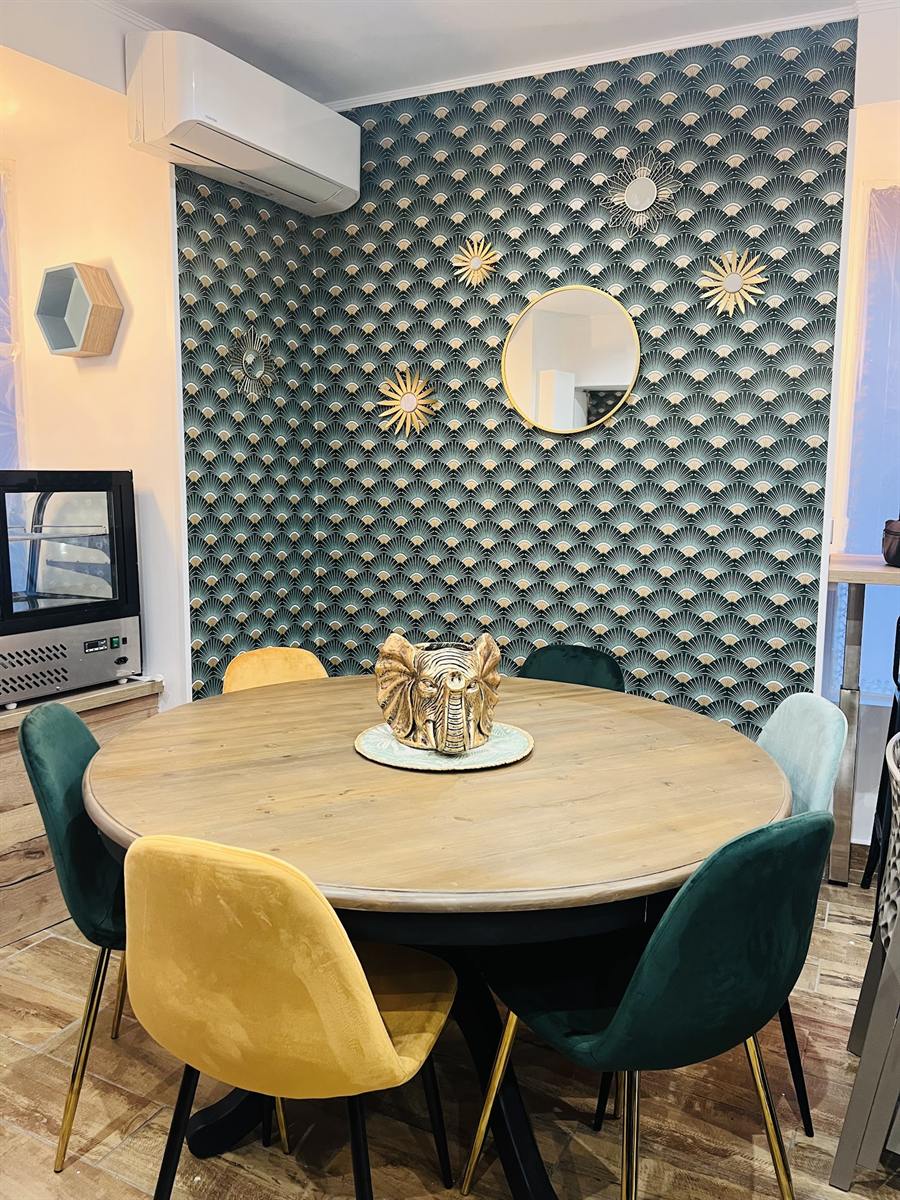 Table ronde en bois et chaises vertes et jaunes devant un papier peint vert à motifs dorés